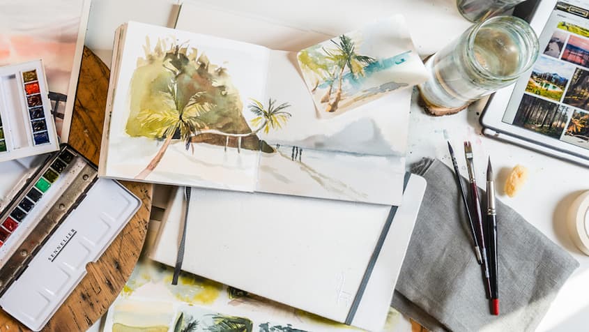 Table recouverte de carnets avec des paysages à l'aquarelle et des pinceaux, palettes, bocaux d'eau
